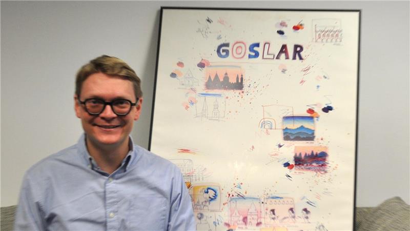 Mann in hellblauem Hemd sitzt auf grauem Sofa vor einem großen, gerahmten Poster mit der Aufschrift 'GOSLAR' und verschiedenen bunten Zeichnungen.