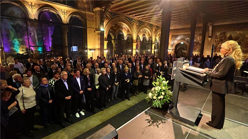 Eine Frau steht an einem Rednerpult in einem großen, prunkvollen Saal vor einem Publikum, das in Reihen steht und ihr zuhört.