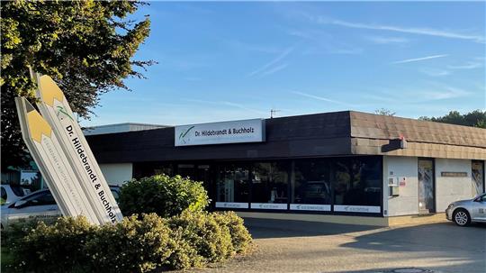 Flacher Geschäftsgebäudekomplex mit großen Fenstern und einem Schild 'Dr. Hildebrandt & Buchholz' davor sowie einer Skulptur aus übergroßen Bleistiften im Vordergrund