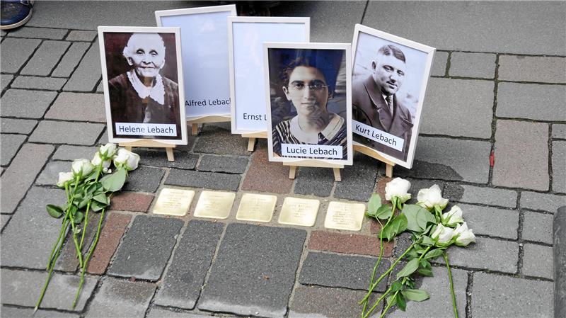 Vier Porträtfotos mit den Namen Helene Lebach, Ernst Lebach, Lucie Lebach und Kurt Lebach auf Pflastersteinen, umgeben von weißen Rosen.