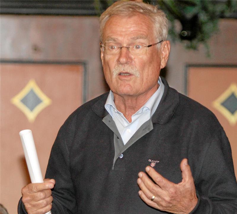 Älterer Mann mit grauem Hemd und schwarzem Pullover hält eine zusammengerollte weiße Papierrolle in der linken Hand und gestikuliert mit der rechten Hand.