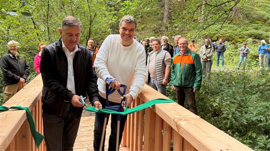 Zwei Männer stehen auf einer Holzbrücke und zerschneiden ein Stoffband.
