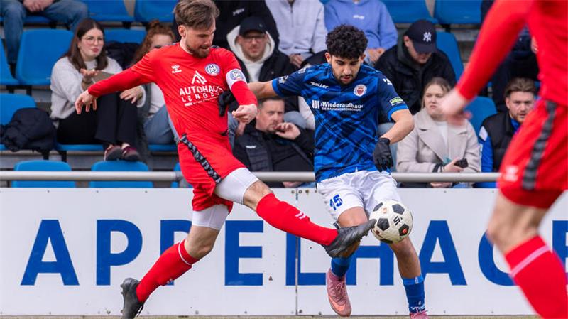 Zwei Fußballspieler in Aktion auf dem Spielfeld, einer in rotem Trikot mit ausgestrecktem Bein, der andere in blauem Trikot mit Ball am Fuß, Zuschauer im Hintergrund auf blauen Sitzen.