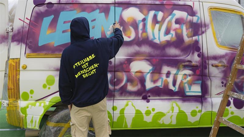 Person mit Kapuzenpullover mit der Aufschrift '#TEILHABE IST MENSCHENRECHT' sprüht Graffiti auf einen weißen Van