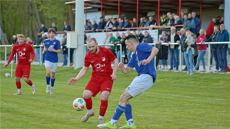 Zwei Fußballspieler in Aktion auf dem Spielfeld, einer in rotem Trikot und einer in blauem Trikot, mit Zuschauern im Hintergrund