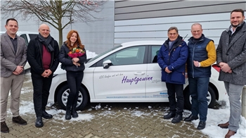 Sechs Personen stehen vor einem weißen Auto mit der Aufschrift 'Hauptgewinn', eine Frau hält einen Blumenstrauß.