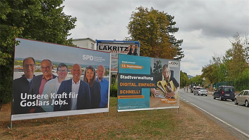 Zwei Wahlplakate an einer Straße mit Bäumen und Autos im Hintergrund, eines von SPD mit mehreren Personen und dem Text 'Unsere Kraft für Goslars Jobs', das andere von CDU mit einer Frau und dem Text 'Stadtverwaltung DIGITAL. EINFACH. SCHNELL!'