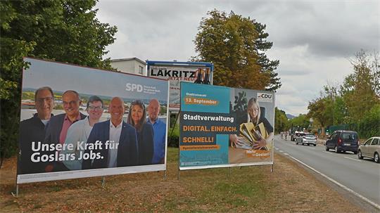 Zwei Wahlplakate an einer Straße mit Bäumen und Autos im Hintergrund, eines von SPD mit mehreren Personen und dem Text 'Unsere Kraft für Goslars Jobs', das andere von CDU mit einer Frau und dem Text 'Stadtverwaltung DIGITAL. EINFACH. SCHNELL!'