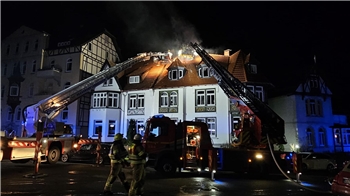 Einsatzkräfte der Feuerwehr und der Polizei stehen vor dem brennenden Haus. Drehleiterin der Feuerwehr sind vor dem Gebäude aufgebaut.