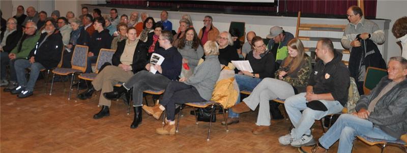 Gruppe von Menschen sitzt in einem Saal auf Stühlen in einer Reihe und unterhält sich teilweise miteinander