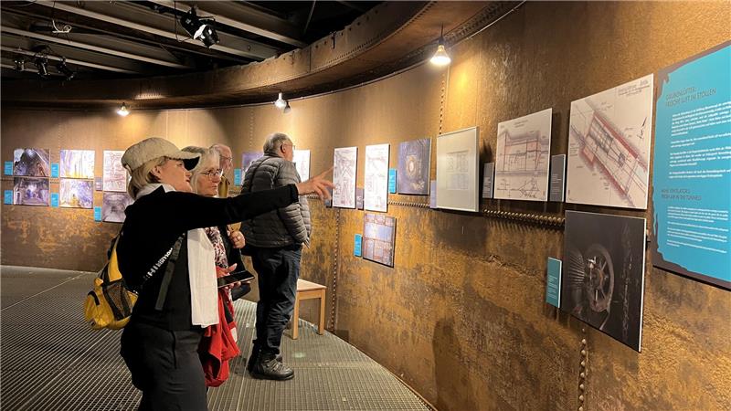 Mehrere Personen betrachten an einer gebogenen Wand hängende Pläne und Zeichnungen in einer Ausstellung.