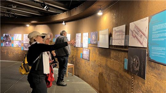 Mehrere Personen betrachten an einer gebogenen Wand hängende Pläne und Zeichnungen in einer Ausstellung.