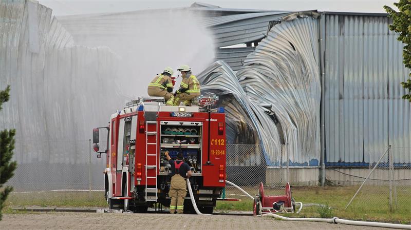 Feuerwehrfahrzeug mit geöffneten Fächern und Feuerwehrleuten löscht Brand an einer großen, beschädigten Industriehalle
