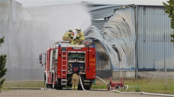 Feuerwehrfahrzeug mit geöffneten Fächern und Feuerwehrleuten löscht Brand an einer großen, beschädigten Industriehalle