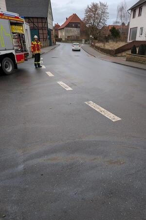 Ölflecken auf einer Fahrbahn, im Hintergrund steht ein Feuerwehrmann an einem Einsatzfahrzeug.