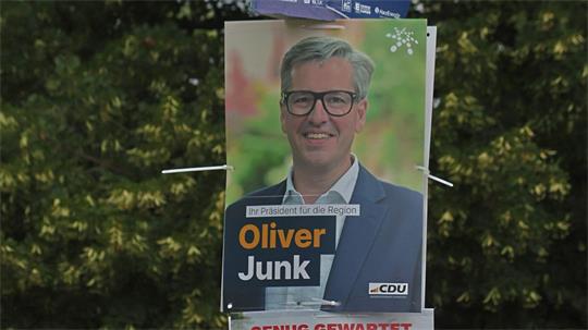 Drei Wahlplakate übereinander an einem Mast mit Porträts von zwei Kandidaten und einem Hinweis auf ein Fest im Hintergrund