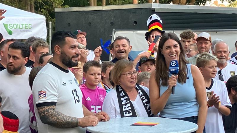 Gruß aus Undavs Heimat Achim nach East Rutherford: Edeltraut Koch (M), erste Trainerin von Deniz Undav, und Aykut Kaldirici (l), Trainer der 1. Herren des TSV Achim und eng befreundet mit Undavs Familie bei einer Schalte mit ARD-Reporterin Anna Klär.
