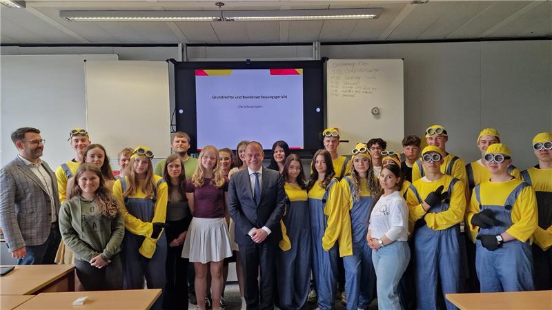 Gruppenfoto von Personen in einem Klassenraum, einige tragen gelbe Minion-Kostüme mit Brillen, andere normale Kleidung, im Hintergrund eine Präsentation und eine Tafel.