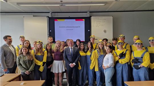 Gruppenfoto von Personen in einem Klassenraum, einige tragen gelbe Minion-Kostüme mit Brillen, andere normale Kleidung, im Hintergrund eine Präsentation und eine Tafel.