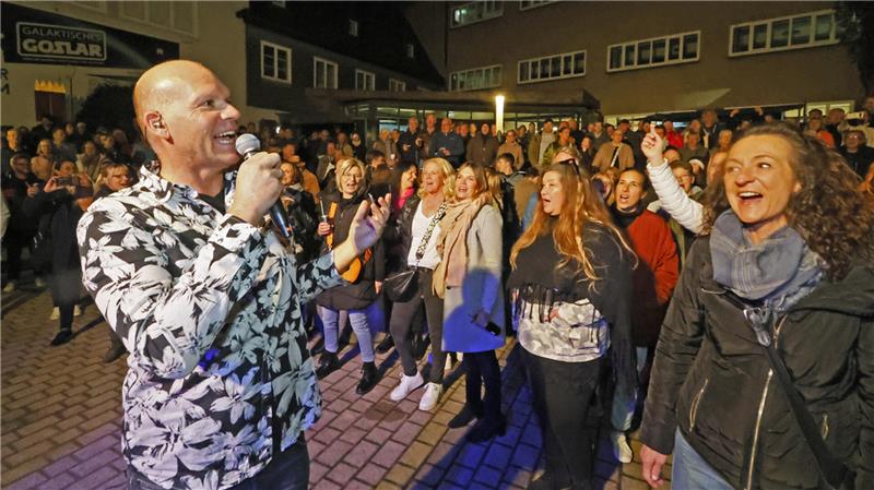 Mann mit Mikrofon singt vor einer großen Menschenmenge auf einem gepflasterten Platz bei Nacht.