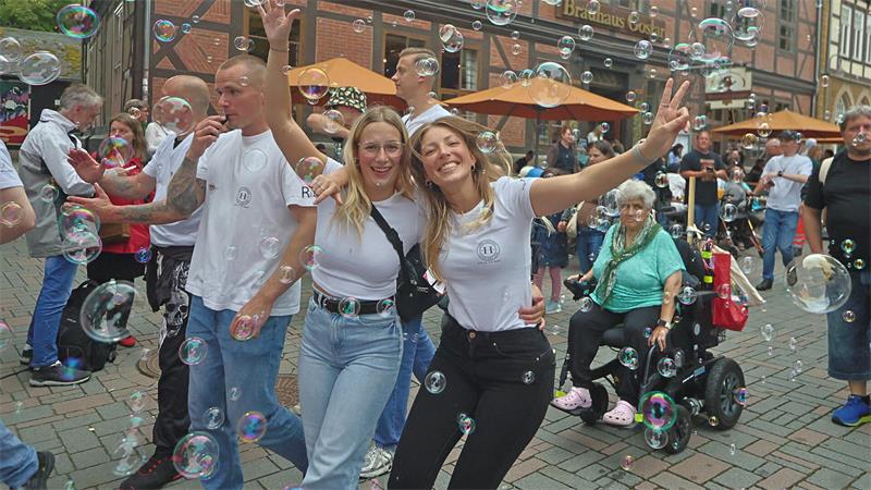 Zwei junge Frauen in weißen T-Shirts und Jeans posieren lachend mit erhobenen Armen zwischen vielen Seifenblasen auf einem belebten Marktplatz