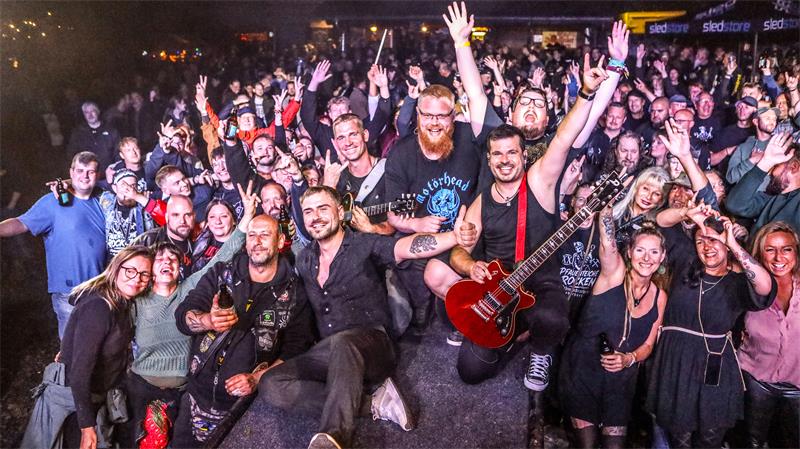 Gruppe von Menschen bei einem Konzert, einige mit erhobenen Händen, ein Mann hält eine rote E-Gitarre auf der Bühne.