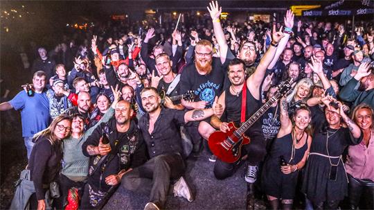 Gruppe von Menschen bei einem Konzert, einige mit erhobenen Händen, ein Mann hält eine rote E-Gitarre auf der Bühne.