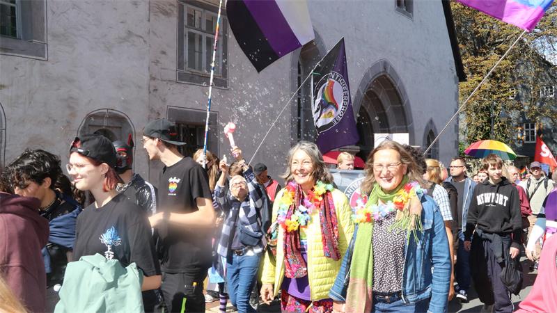 Menschenmenge bei einem Straßenumzug mit bunten Fahnen und Blumenketten, zwei Frauen im Vordergrund lächeln.