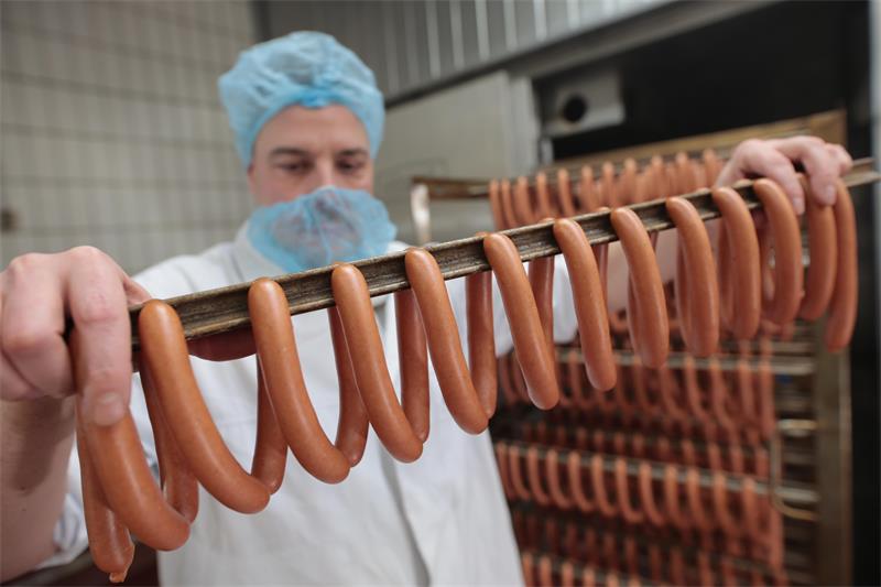 Ein mit Kittel, Mund- und Kopfschutz ausgestatteter Mann hält eine Stange mit Bockwürstchen.