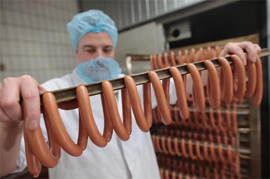 Ein mit Kittel, Mund- und Kopfschutz ausgestatteter Mann hält eine Stange mit Bockwürstchen.