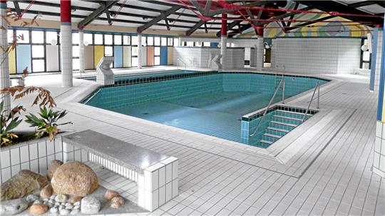 Innenaufnahme eines leeren Schwimmbeckens in einer Halle mit weißen Fliesen und großen Fenstern