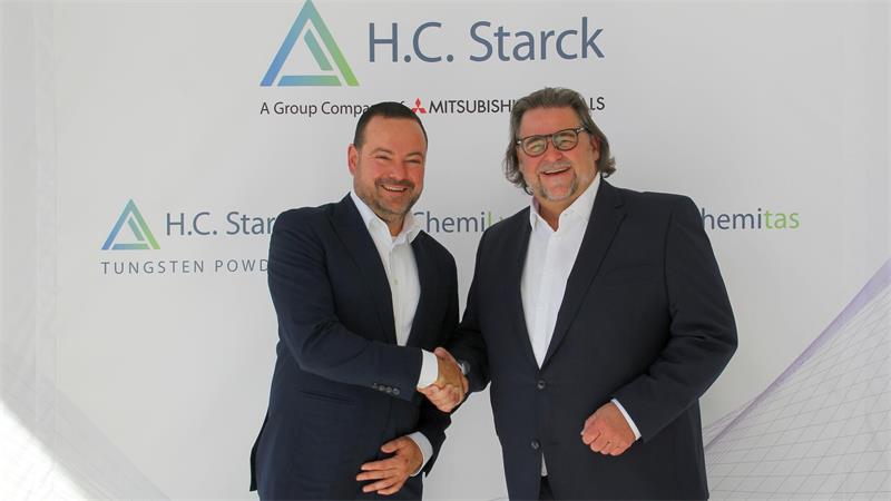 Zwei Männer in Anzügen schütteln sich vor einer Wand mit dem Logo von H.C. Starck die Hand