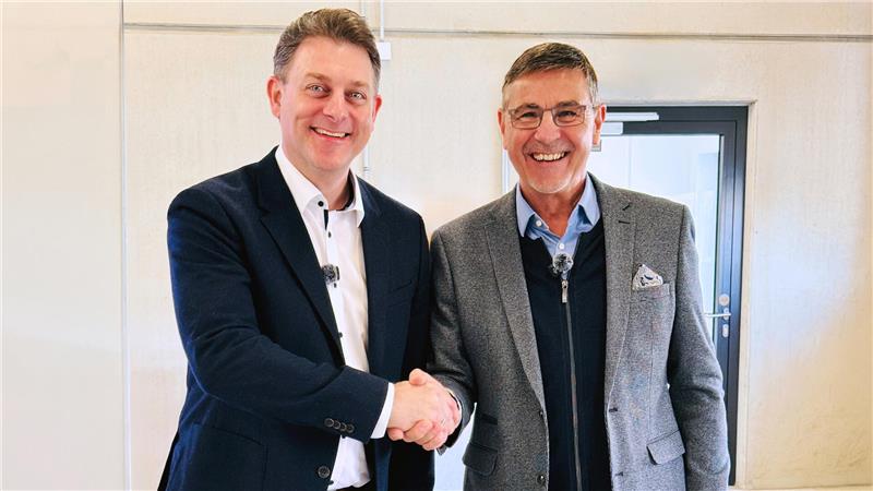 Zwei Männer in Anzügen schütteln sich die Hand vor einer weißen Wand mit Tür