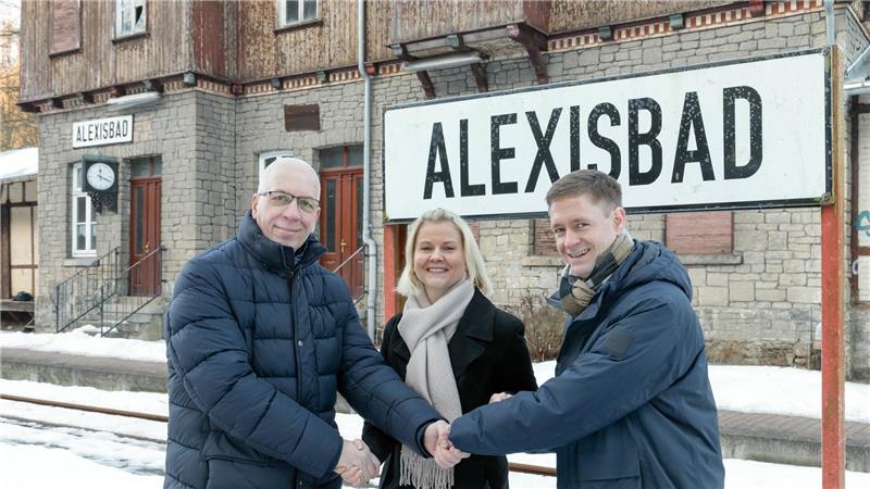 Drei Personen in Winterkleidung stehen vor einem Bahnhofsgebäude mit den Schildern ALEXISBAD' und schütteln sich die Hände.