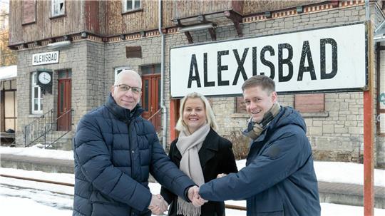 Drei Personen in Winterkleidung stehen vor einem Bahnhofsgebäude mit den Schildern ALEXISBAD' und schütteln sich die Hände.