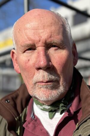 Postbank-Schließung: Das sagen Kunden, die FDP will eine Petition Älterer Mann mit weißem Haar trägt braune Jacke, weißes T-Shirt und grünes Halstuch vor unscharfem Hintergrund mit Stahlträgern