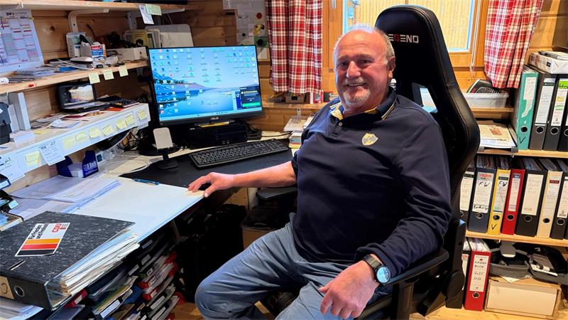 Ein Mann in einem dunklen Pullover und einer hellen Jeans sitzt auf einem Bürostühl vor einem Schreibtisch mit einem Computer.