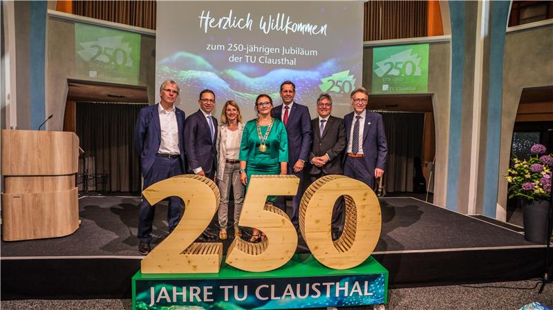 Sieben Männer und Frauen stehen auf einer Bühne. Vor ihnen ist eine aus Holz geschnitzte "250" aufgestellt.