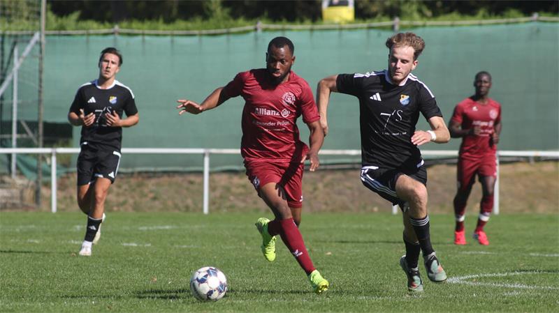 Zwei Fußballspieler in Aktion auf dem Spielfeld, einer in rotem Trikot mit Ball, der andere in schwarzem Trikot läuft neben ihm