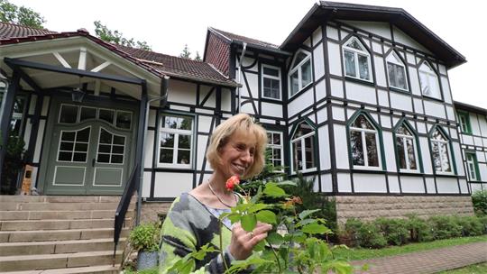 Eine Frau hält die Blüte einen Rosenstrauchs in der Hand, im Hintergrund ein Fachwerkhaus.