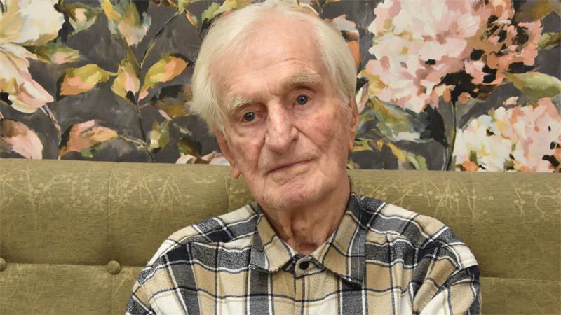 Älterer Mann mit kariertem Hemd sitzt mit verschränkten Armen vor einer floral gemusterten Wand.