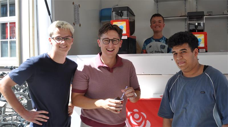 Vier junge Männer stehenvor einem Eiswagen, drei vorne und einer hinten an einem Tresen, zwie der vorderen tragen eine Brille 