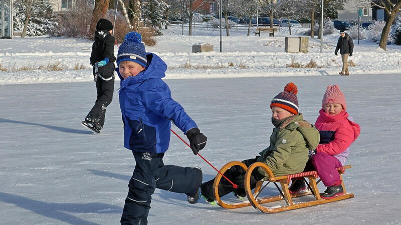 Zwei Kinder sitzen auf einem Schlitten auf einer zugefrorenen Fläche, ein Kind zieht den Schlitten, weitere Personen im Hintergrund auf dem Eis.