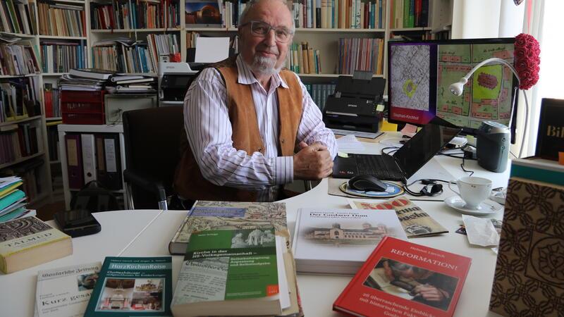 Älterer Mann mit Brille und brauner Weste sitzt an einem Tisch mit mehreren Büchern in einem Raum voller Bücherregale.