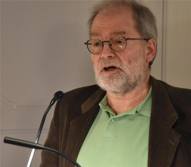 Mann mit grauem Haar, Brille und grünem Poloshirt unter braunem Sakko vor Mikrofon stehend