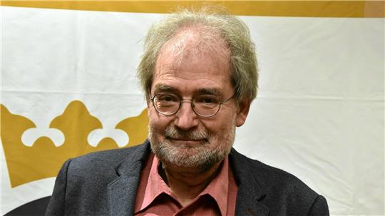 Ein älterer Mann mit grauem Haar trägt ein dunkelgraues Sakko und ein rotes Hemd vor einem weißen Hintergrund mit einem goldfarbenen Kronensymbol.