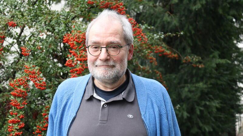 Älterer Mann mit grauem Haar trägt ein graues Poloshirt und eine blaue Strickjacke vor einem Strauch mit roten Beeren