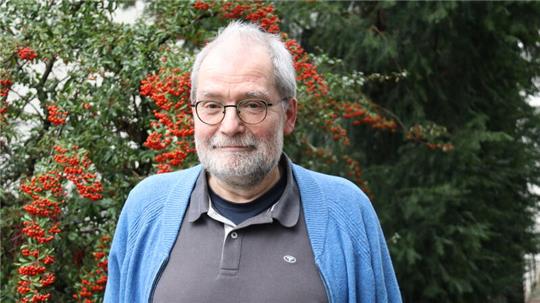 Älterer Mann mit grauem Haar trägt ein graues Poloshirt und eine blaue Strickjacke vor einem Strauch mit roten Beeren