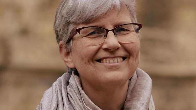 Eine Frau mit kurzen grauen Haaren trägt einen hellen Schal und eine rot-braune Jacke vor unscharfem Hintergrund.