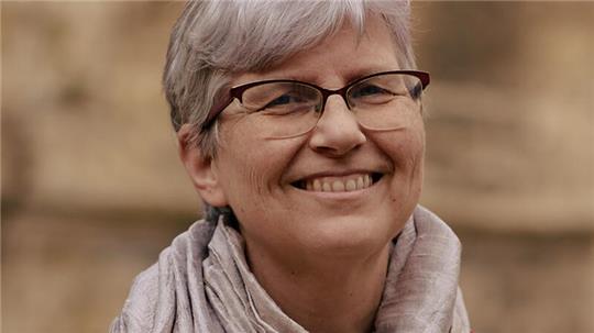 Eine Frau mit kurzen grauen Haaren trägt einen hellen Schal und eine rot-braune Jacke vor unscharfem Hintergrund.